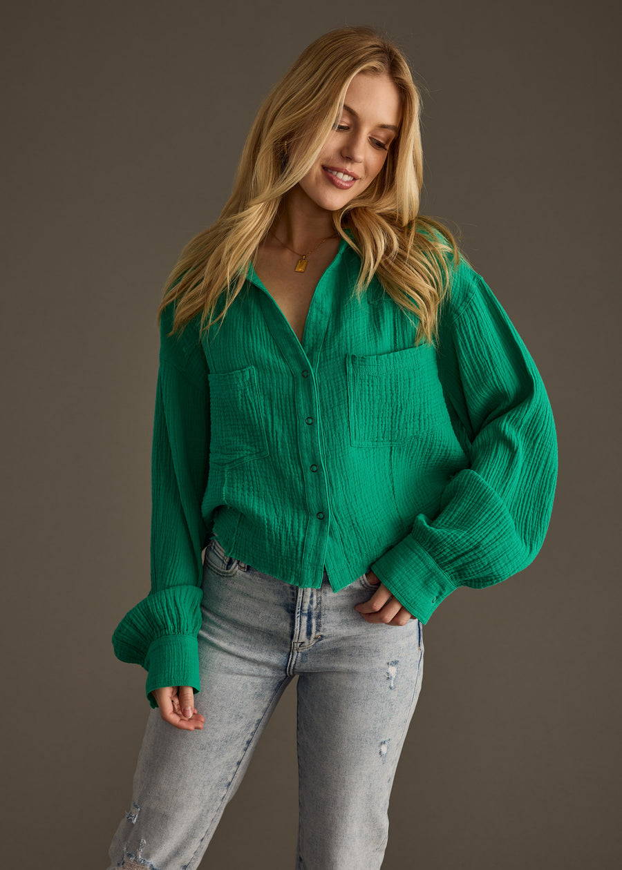A woman with long blonde hair wears the panacheaccessories Wild Heart Button Up in green, styled oversized with light blue ripped jeans, standing against a plain brown background and smiling while looking slightly down.
