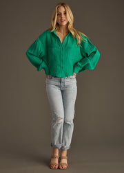 A woman with long blonde hair stands against a plain background wearing the panacheaccessories Wild Heart Button Up - Green, paired with light blue ripped jeans and tan heeled sandals. Her hands rest on her hips.