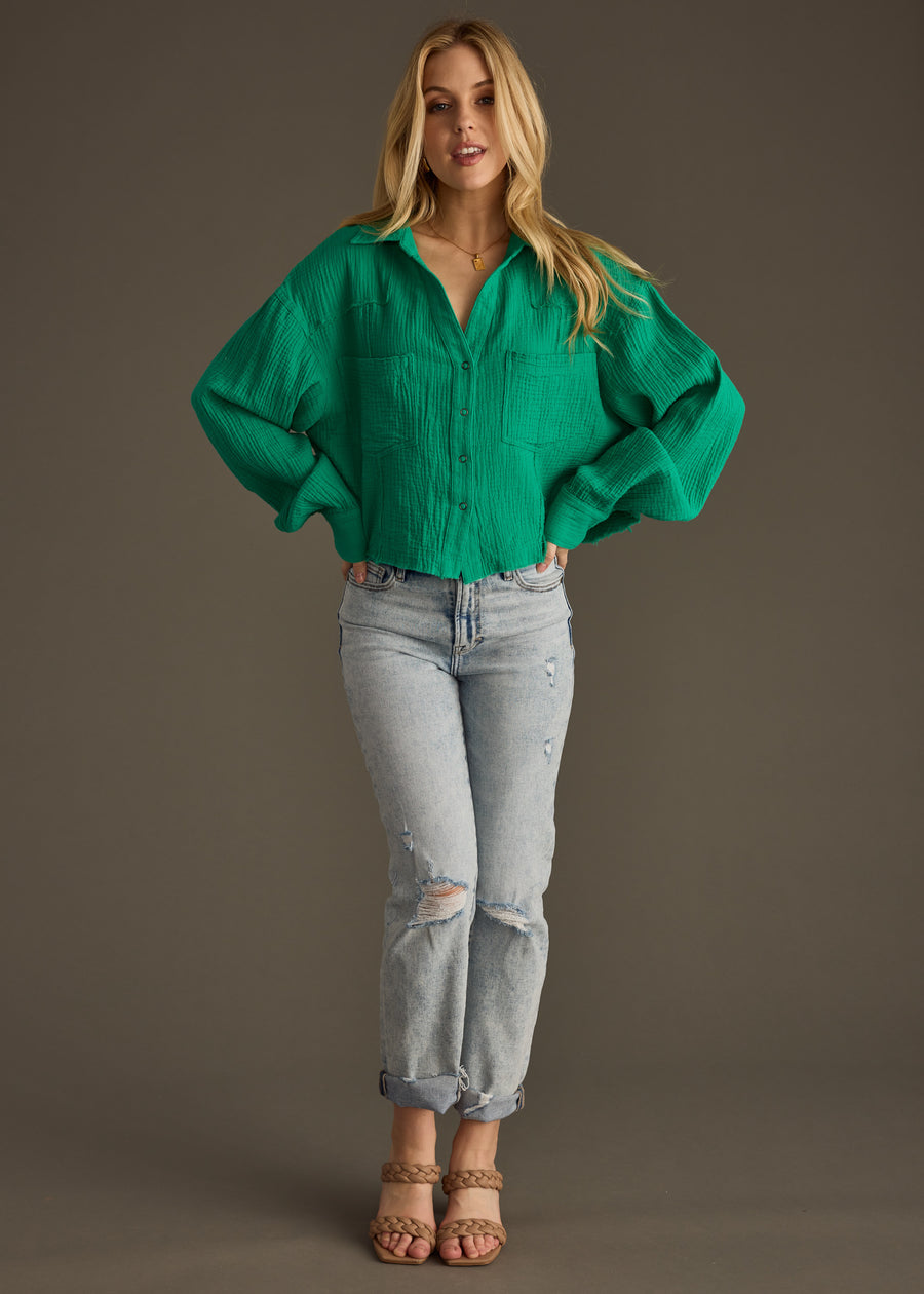 A woman with long blonde hair stands against a plain background wearing the panacheaccessories Wild Heart Button Up - Green, paired with light blue ripped jeans and tan heeled sandals. Her hands rest on her hips.
