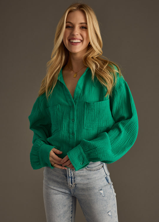 A smiling woman with long blonde hair wears the Wild Heart Button Up - Green from panacheaccessories, styled in an oversized fit with light-wash ripped jeans, standing against a plain dark background.
