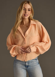A woman with long blonde hair wears the Wild Heart Button Up – Orange from panacheaccessories and light blue jeans, standing against a plain brown background and smiling slightly at the camera.