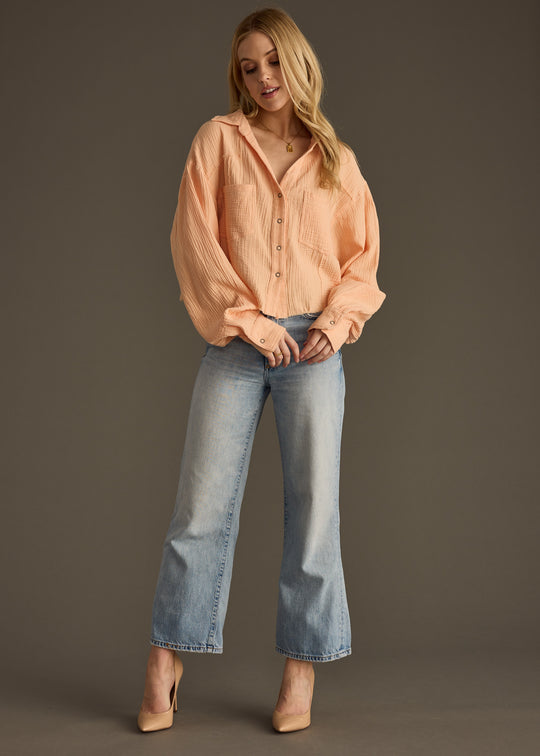 A woman stands against a neutral background, looking down with hands clasped, wearing the panacheaccessories Wild Heart Button Up in orange, styled oversized with light blue jeans and nude heels.