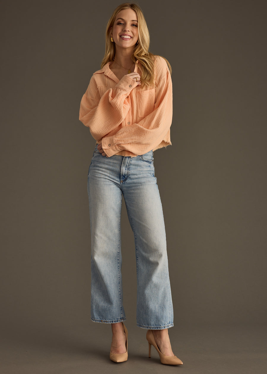 A smiling woman with long blonde hair poses against a plain background wearing the Wild Heart Button Up - Orange from panacheaccessories, styled oversized, paired with light blue high-waisted jeans and beige heels. One hand is raised toward her collar.