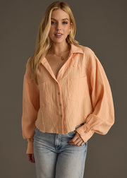 A woman with long blonde hair wears the Wild Heart Button Up - Orange by panacheaccessories, an oversized light orange shirt with large front pockets, paired with light blue jeans, standing against a plain dark background.