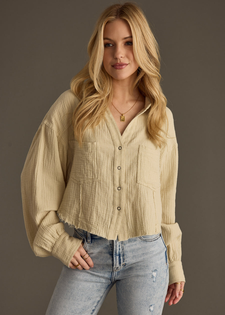 A woman with long blonde hair smiles softly at the camera, wearing the Wild Heart Button Up - Khaki from panacheaccessories and light blue cropped ripped jeans, standing against a plain gray background.