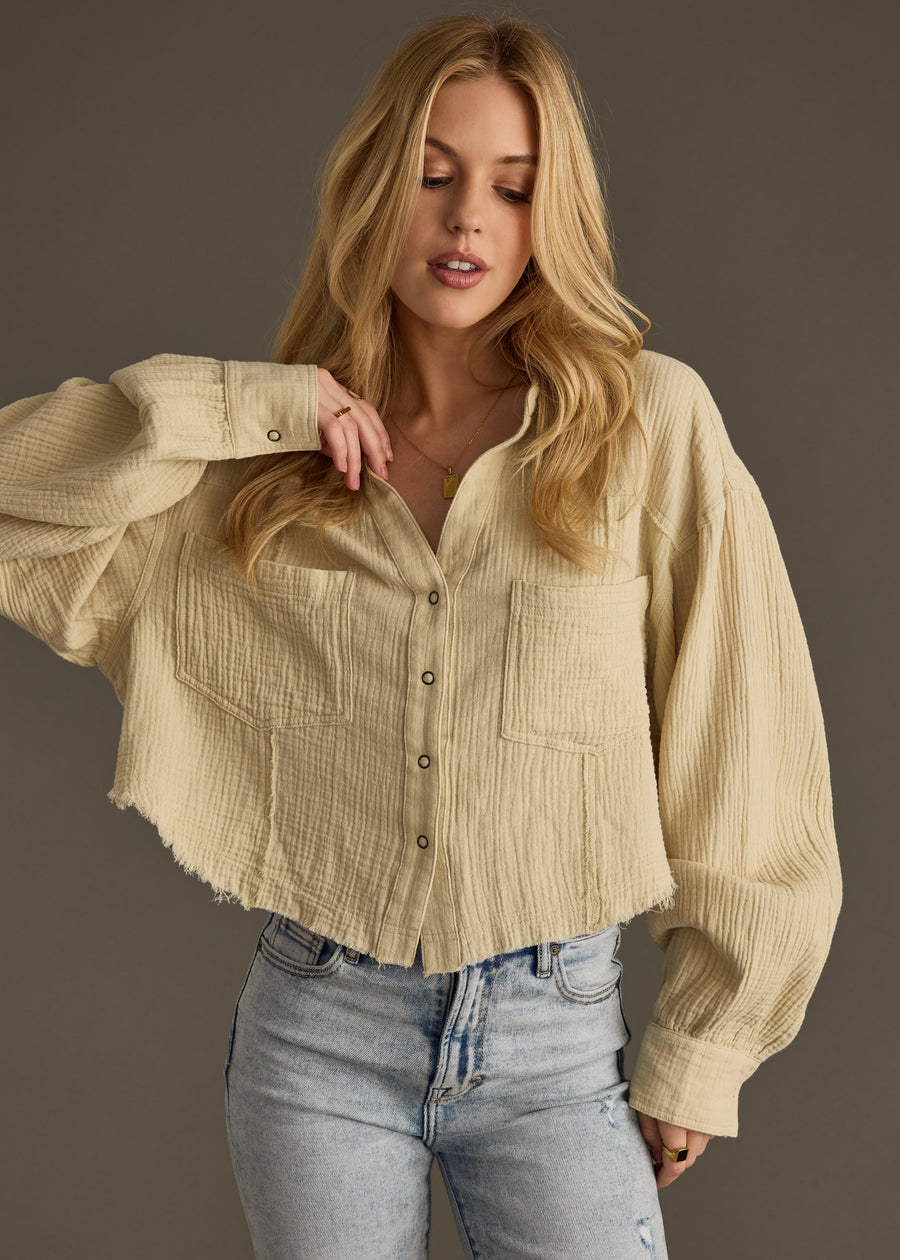 A woman with long blonde hair models the panacheaccessories Wild Heart Button Up - Khaki, featuring frayed edges and a cropped hemline, paired with light blue high-waisted jeans against a plain gray background.