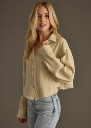 A woman with long blonde hair models the panacheaccessories Wild Heart Button Up in khaki—a frayed edge, oversized shirt—paired with light blue jeans. She poses against a dark background, gazing at the camera with a relaxed look.