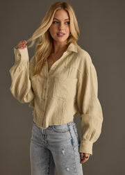 A woman with long blonde hair wears the Wild Heart Button Up - Khaki by panacheaccessories and light blue ripped jeans, standing against a dark background while touching her hair and looking to the side.