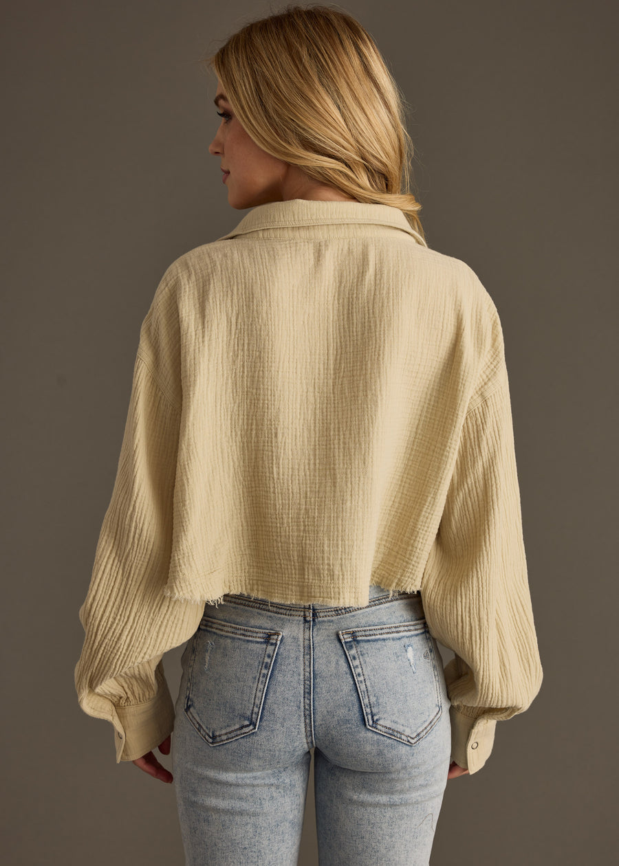 A woman with long blonde hair wears the Wild Heart Button Up in khaki by panacheaccessories, featuring a cropped hemline and loose sleeves, paired with light-wash high-waisted jeans, facing away against a plain dark background.