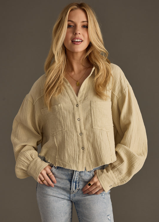 A woman with long blonde hair wears the panacheaccessories Wild Heart Button Up in khaki, featuring an oversized boxy fit and large sleeves, paired with light blue jeans. She stands against a dark background, smiling slightly with her hands in her pockets.