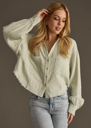A woman stands against a plain background wearing the panacheaccessories Wild Heart Button Up in sage and light blue high-waisted jeans, her left hand touching her hair as she gazes downward.
