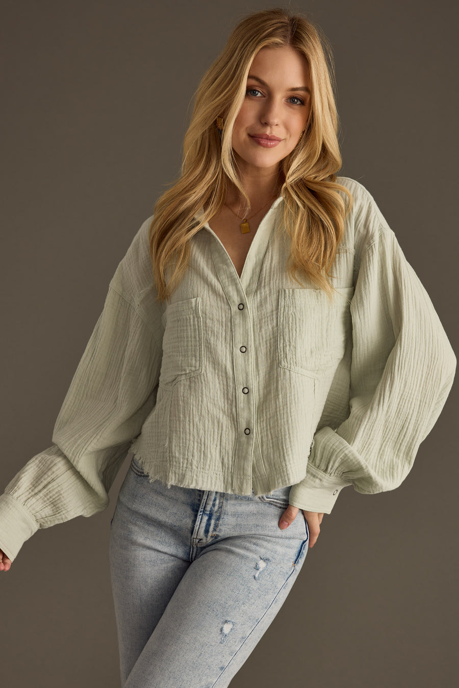 A young woman with long blonde hair models the panacheaccessories Wild Heart Button Up in sage, featuring an oversized boxy fit and two chest pockets, paired with light blue jeans against a dark background.