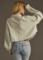 A woman with long blonde hair wears the panacheaccessories Wild Heart Button Up in sage, styled with light blue high-waisted jeans. She stands with her back to the camera, raising one hand to her forehead.