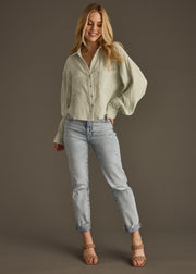 A woman smiles, wearing the panacheaccessories Wild Heart Button Up in sage with an oversized fit, light blue cuffed jeans, and tan strappy heels, posing against a plain brown background.