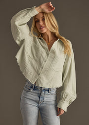 A woman with long blonde hair poses with one arm raised against a gray background, wearing the panacheaccessories Wild Heart Button Up in sage and light blue jeans.