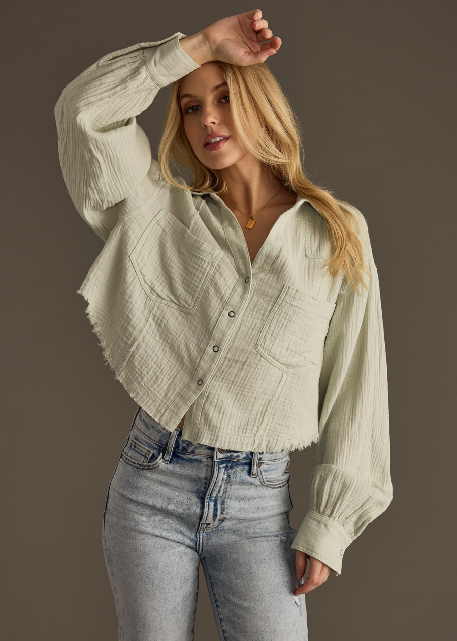 A woman with long blonde hair poses with one arm raised against a gray background, wearing the panacheaccessories Wild Heart Button Up in sage and light blue jeans.