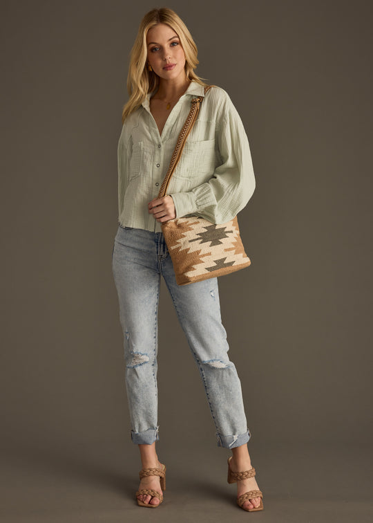 A woman stands against a plain background wearing the Wild Heart Button Up - Sage by panacheaccessories, light blue ripped jeans, heeled sandals, and a woven geometric crossbody bag.