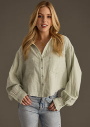 A woman with long blonde hair, wearing the panacheaccessories Wild Heart Button Up in sage and light blue jeans, stands against a plain gray background, smiling at the camera with her hands in her pockets.
