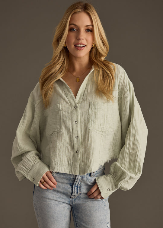 A woman with long blonde hair, wearing the panacheaccessories Wild Heart Button Up in sage and light blue jeans, stands against a plain gray background, smiling at the camera with her hands in her pockets.