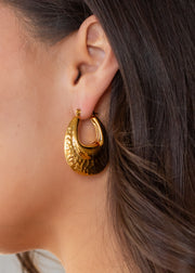 Close-up of a womans ear wearing panacheaccessories Luxe Gold Hammered Bubble Hoops. These shiny 18K gold, teardrop-shaped hoops are waterproof, combining elegance and durability. Her dark brown hair is tucked behind her ear.