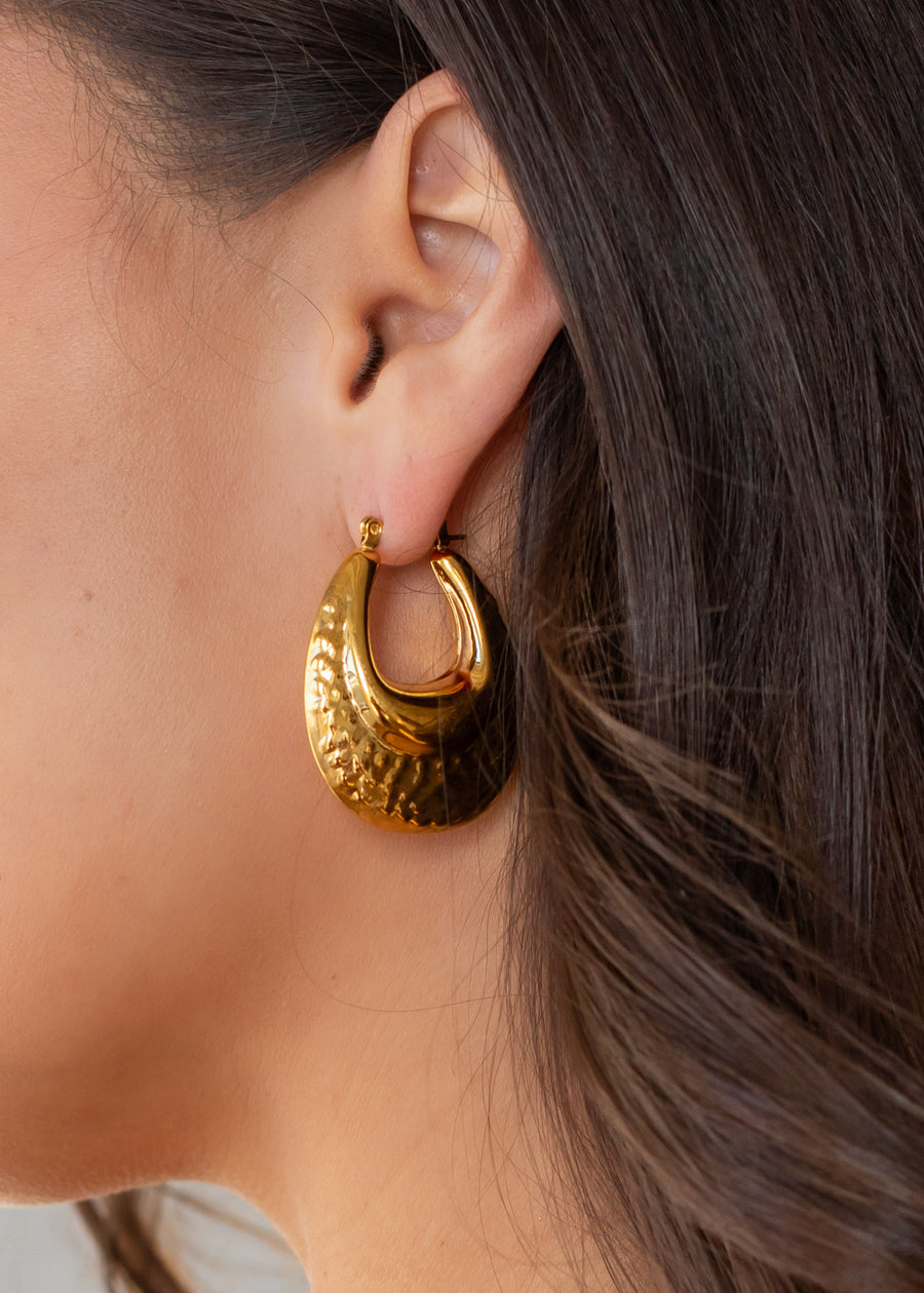 Close-up of a womans ear wearing panacheaccessories Luxe Gold Hammered Bubble Hoops. These shiny 18K gold, teardrop-shaped hoops are waterproof, combining elegance and durability. Her dark brown hair is tucked behind her ear.