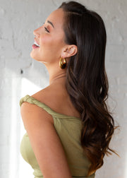 A woman with long, wavy brown hair wearing a green off-shoulder top and panacheaccessories Luxe Gold Hammered Bubble Hoops smiles, looking left in front of a white brick wall.