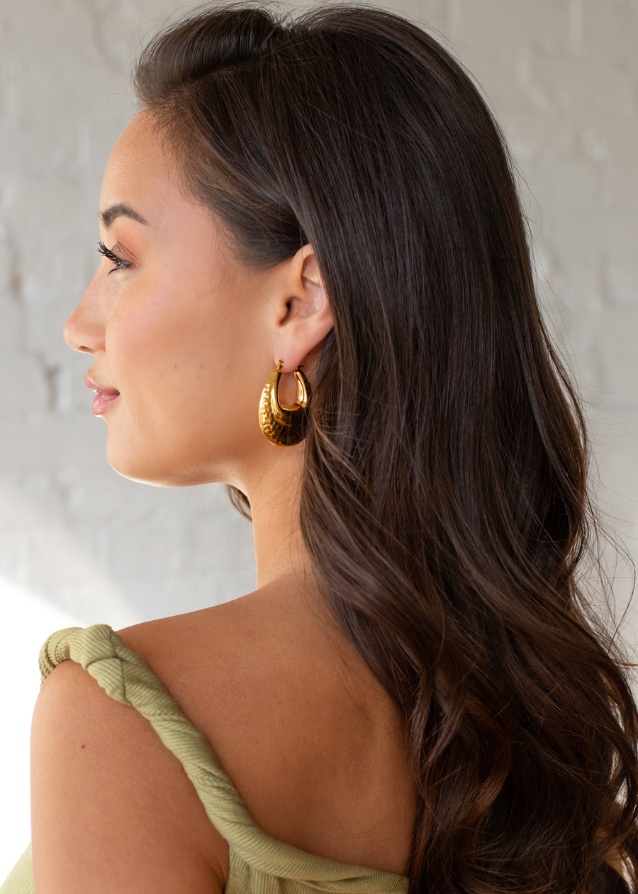 A woman with long, wavy brown hair in a light green top wears panacheaccessories’ Luxe Gold Hammered Bubble Hoops, pictured in profile against a light-colored brick wall.