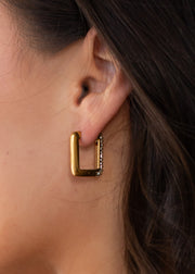 Close-up of a womans ear adorned with panacheaccessories Luxe Gold & Rhinestone Squared Hoop Earrings—rectangular, gold, and accented with sparkly rhinestones. These elegant waterproof hoops shine against her dark hair.