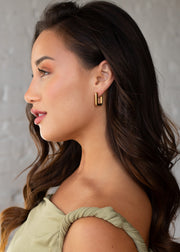 A woman with long, wavy brown hair wears a green twisted-strap top and panacheaccessories’ Luxe Gold & Rhinestone Squared Hoop Earrings. She is shown in profile against a light textured background, wearing natural makeup and a slight smile.