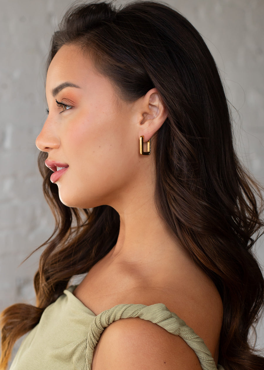 A woman with long, wavy brown hair wears a green twisted-strap top and panacheaccessories’ Luxe Gold & Rhinestone Squared Hoop Earrings. She is shown in profile against a light textured background, wearing natural makeup and a slight smile.