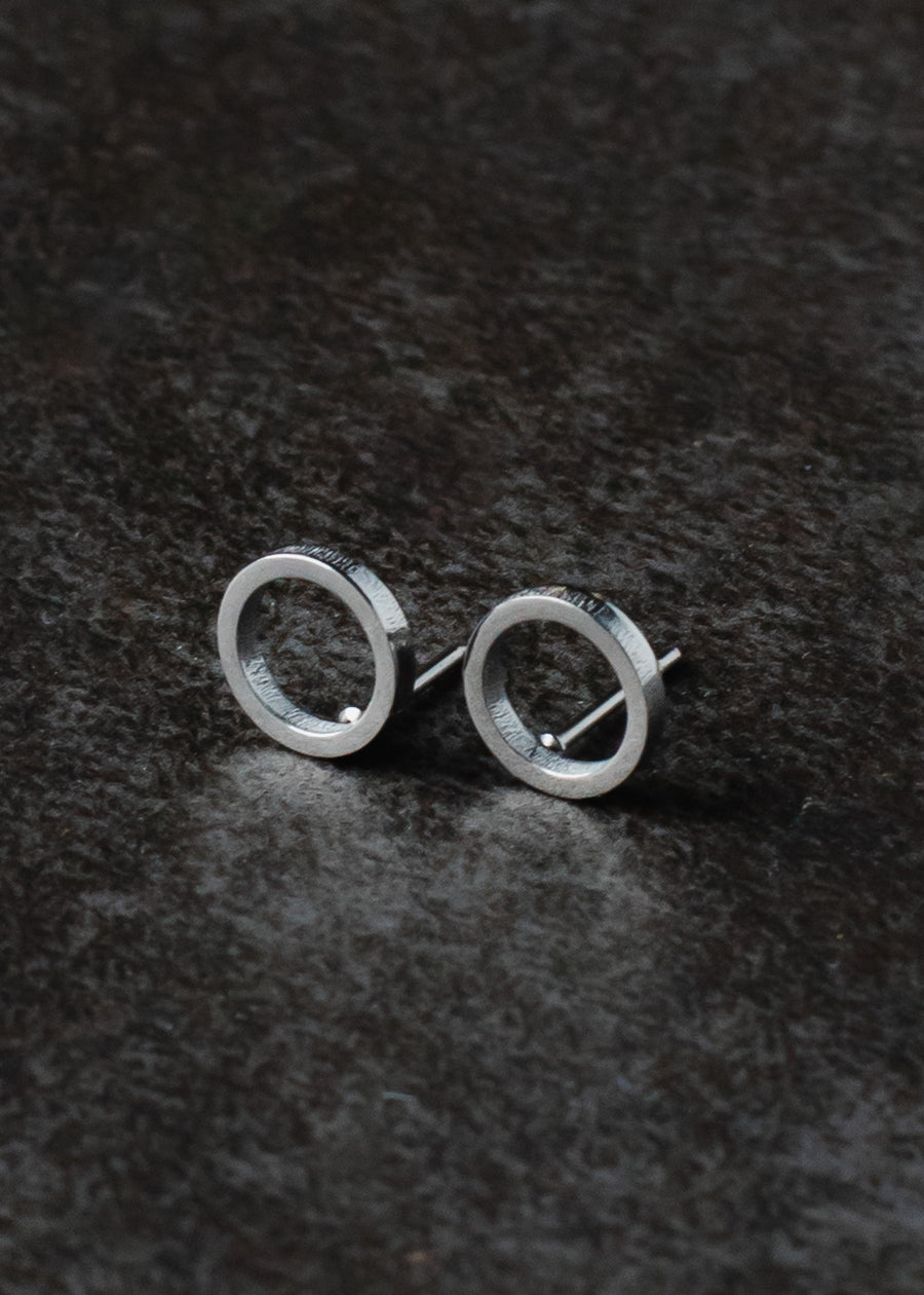 The Begin Again Stud Earrings - Silver by panacheaccessories are minimalist open circle studs, crafted from lead and nickel free stainless steel, shown on a dark textured surface.
