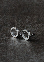 The Begin Again Stud Earrings - Silver by panacheaccessories, minimalist open circle studs that are lead and nickel free, are displayed on a dark, textured surface.