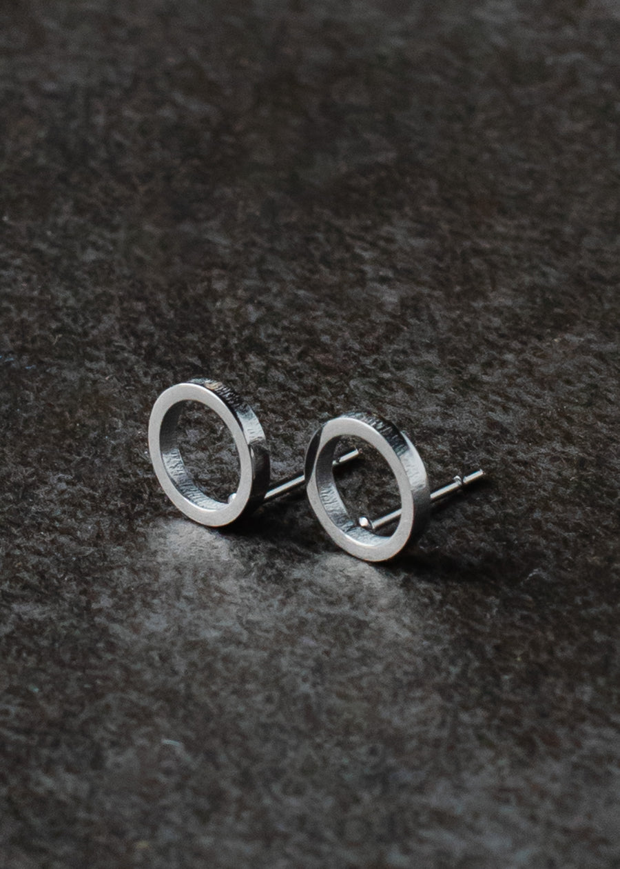 The Begin Again Stud Earrings - Silver by panacheaccessories, minimalist open circle studs that are lead and nickel free, are displayed on a dark, textured surface.