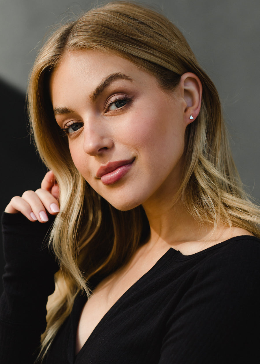A young woman with long blonde hair and light makeup smiles softly at the camera, wearing a black top and panacheaccessories Scout Stud Earrings in gold. She holds her hand near her face against a blurred, neutral background.