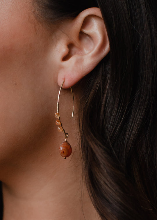 Close-up of a womans ear wearing panacheaccessories Summer Romance Earrings—long gold hoops with small orange beads and a large round stone. These statement earrings are lead and nickel free, ideal for sensitive skin.