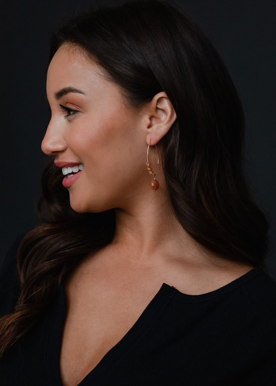 A woman with long dark hair smiles in profile against a dark background, wearing a black top and panacheaccessories’ Summer Romance Earrings—elegant gold earrings featuring a small orange pendant.