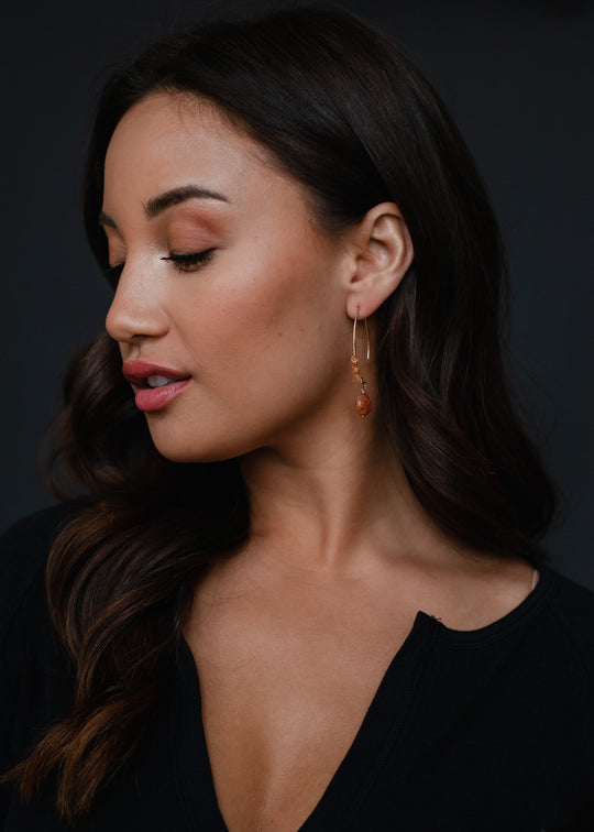 A woman with long, dark hair and natural makeup looks to the side, showcasing panacheaccessories’ Summer Romance Earrings—lead and nickel free gold detail with a small brown stone. She wears a black top against a dark background.