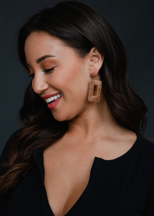 A woman with long dark hair smiles while looking down, wearing a black top and large Meredith Wicker Earrings by panacheaccessories, which are lead and nickel free, against a dark background.