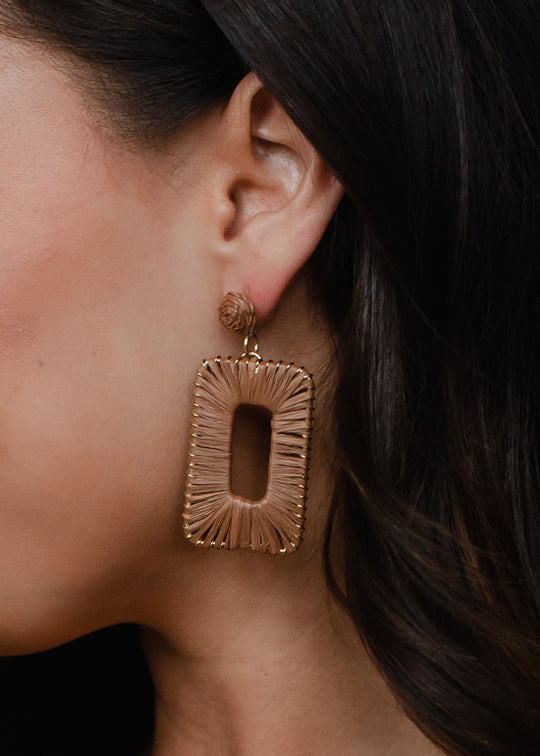 A close-up shows a woman with dark hair pulled back, wearing panacheaccessories Meredith Wicker Earrings—lead & nickel free rectangles with gold frames and light brown woven material, plus a matching round stud.