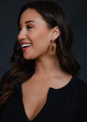 A woman with long dark hair smiles while looking to the side, wearing a black top and panacheaccessories Meredith Wicker Earrings against a dark background.