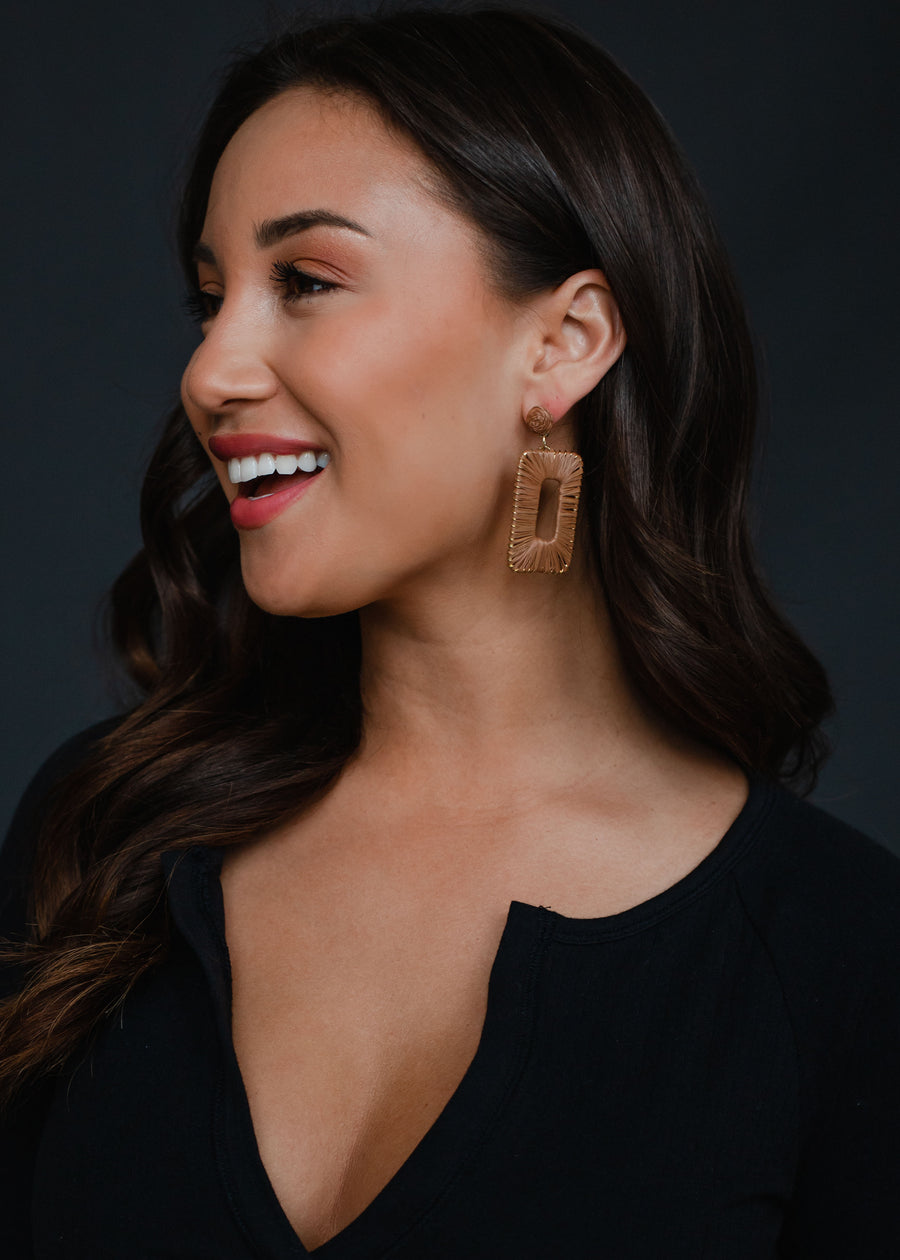 A woman with long dark hair smiles while looking to the side, wearing a black top and panacheaccessories Meredith Wicker Earrings against a dark background.
