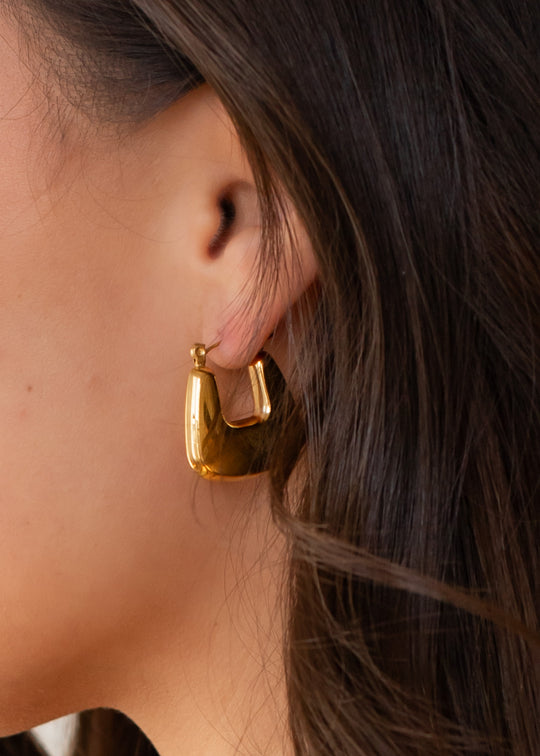 Close-up of a womans ear with long brown hair, wearing panacheaccessories Luxe Gold Hollow Squared Hoops—chunky, modern earrings crafted from durable stainless steel for stylish everyday elegance.