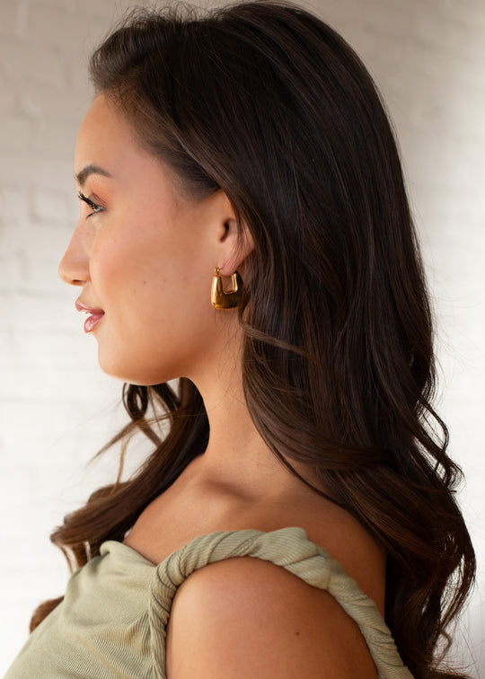 A woman with long, wavy dark hair is shown in profile against a white brick background, wearing panacheaccessories Luxe Gold Hollow Squared Hoops and a light green off-the-shoulder top.
