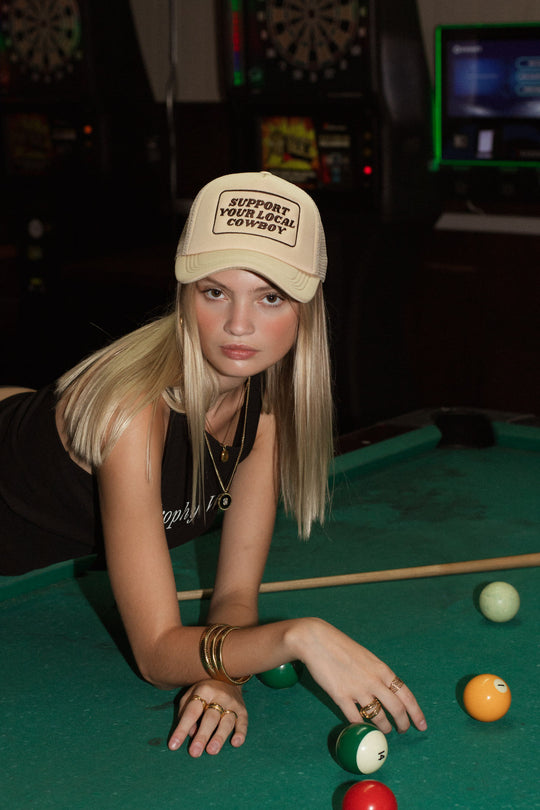 A young woman with long blonde hair leans on a pool table wearing the Panache Apparel Collection’s Support Your Local Cowboy Trucker Hat, a tan cap with an adjustable snapback, paired with black clothing and jewelry among pool balls and cues.