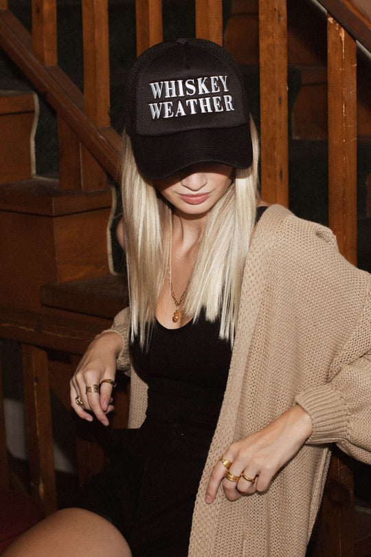A woman with long blonde hair sits on wooden stairs, her face partly hidden by the Panache Apparel Collection’s Whiskey Weather Trucker Hat in black, paired with a black top and beige cardigan.