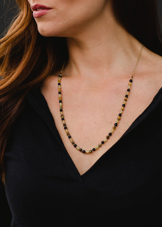 A woman with long brown hair wears the panacheaccessories Annette Beaded Necklace, a 22 lead- and nickel-free piece featuring alternating gold and dark beads, paired with a black V-neck top. Image shows her lower face, neck, and upper chest.