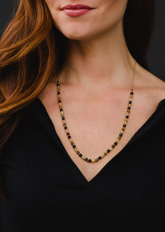 A woman wears the Annette Beaded Necklace by panacheaccessories—a delicate, 22-inch gold and dark-beaded v-neck piece thats lead and nickel free—shown from the shoulders up in a black top against a dark background.