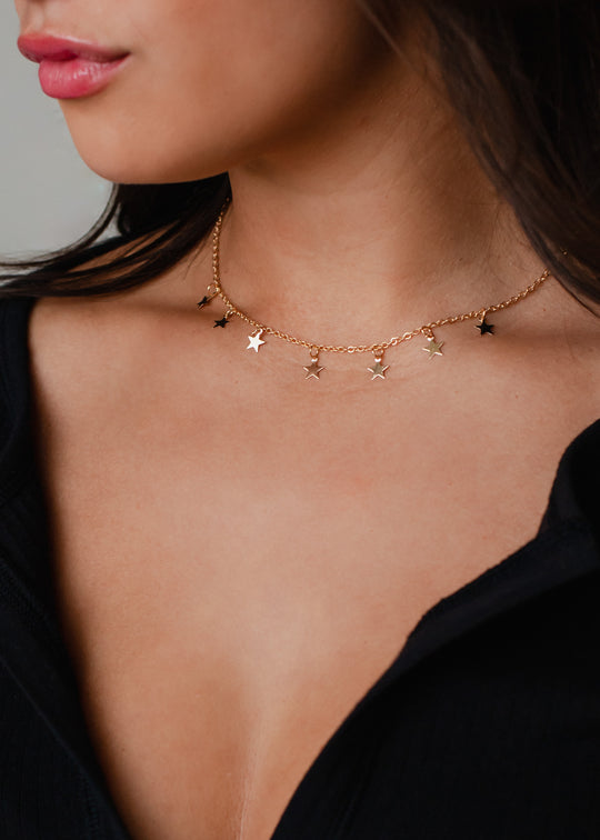 A woman wears the Davina Necklace - Gold by panacheaccessories, a lead and nickel free choker with delicate star charms, paired with a black top. The image focuses on her neck and collarbone.