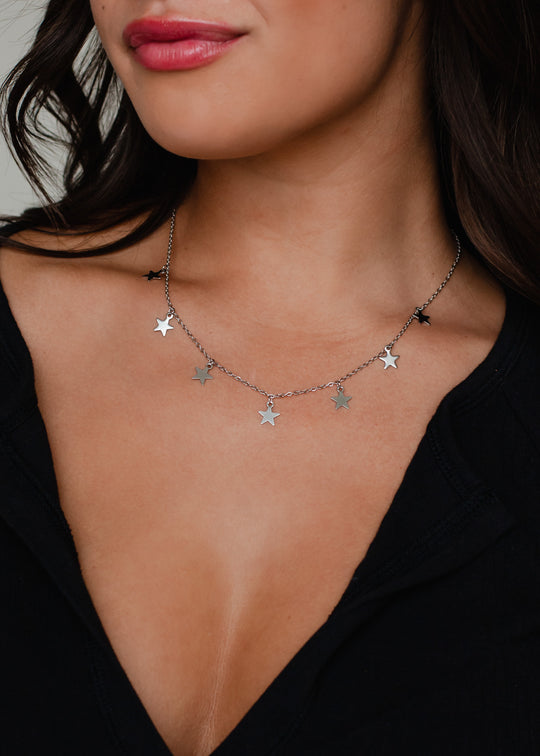 A woman in a black top wears the panacheaccessories Big Dipper Necklace, a delicate, lead and nickel free silver piece with star-shaped pendants. Her wavy dark hair frames her face and she has light pink lipstick.