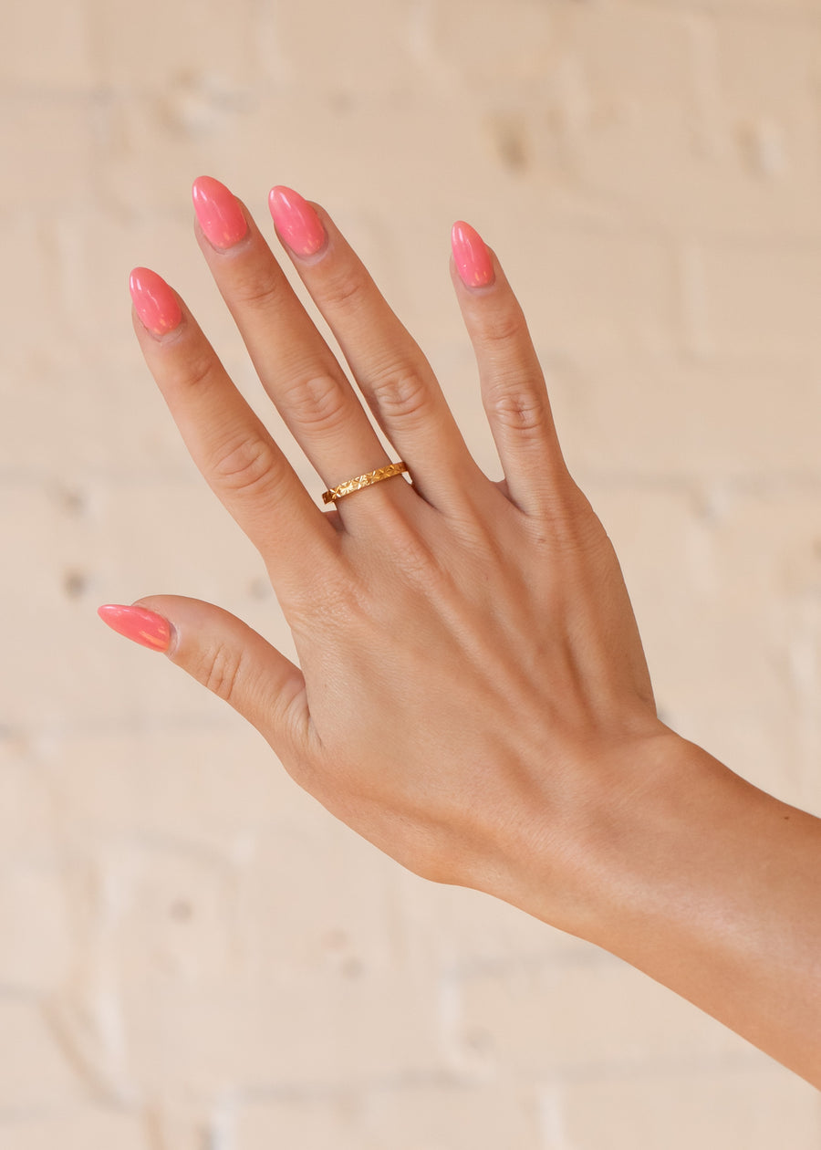 A hand with long, glossy pink nails wears the panacheaccessories Luxe Gold Starburst Stamped Ring on the ring finger, set against a light beige, textured background.